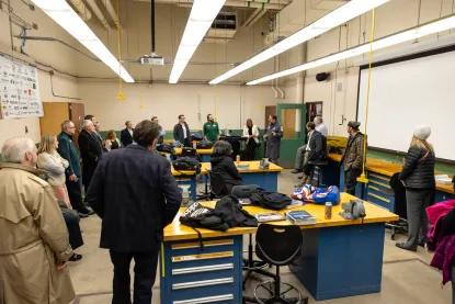 A group of people stand in a classroom or lab, listening to a speaker.