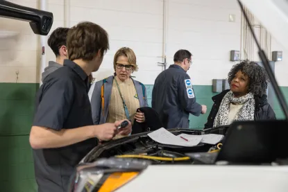Two auto technicians talk with two women beside a vehicle with its hood open.