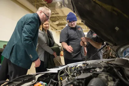 Three adults examine a car engine in a workshop.