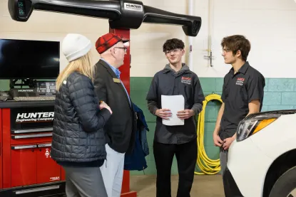 Two students in dark uniforms speak with a man and a woman, inside an auto shop with equipment and a white vehicle visible.