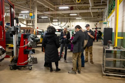 A group of people stands and talks inside an automotive workshop, surrounded by cars, tools, and equipment.