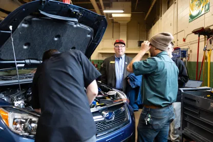 Three people work on a car engine in a garage while a man in a suit jacket observes.