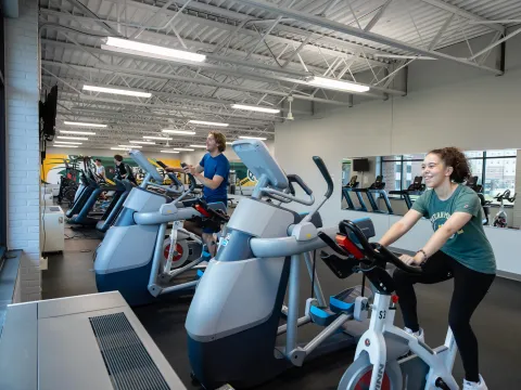 The Wildcat Fit Zone is a small yet spacious gym. Two students use cardio machines; a smiling woman pedals a stationary bike while another exercises on an elliptical.