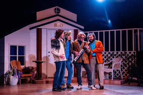 A theater stage set up as a house exterior, with four actors standing together in front of the entrance. One actor holds a small keyboard, while patio furniture, potted flowers, and a lattice fence complete the set under colorful stage lighting.