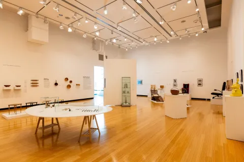 Interior art gallery with white walls, track lighting, and wooden floors displaying furniture, ceramics, sculptures, and other artworks on tables, shelves, and pedestals.