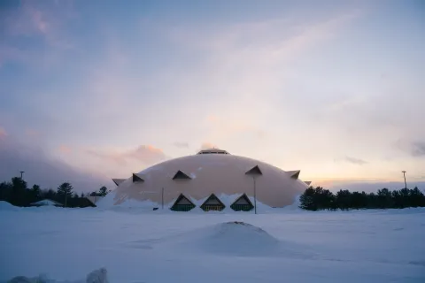 A large white wooden dome with triangular windows sits in a snowy field at dusk, beneath a pink-and-blue sky.