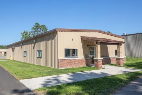 A single-story building with beige metal siding and red brick lower exterior. A concrete walkway leads to the entrance, surrounded by grass.