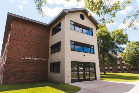 Exterior view of Hunt Hall, a modern university building with red brick wings and a pale stone entrance stands beside green lawns and walkways; bicycles sit in the sunshine under a blue sky.