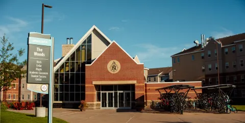 The Lodge is a red-brick building with large windows. Crowded bike racks sits in bright sunshine; a campus sign reads “The Woods.”