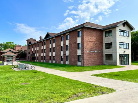 Exterior view of Magers Hall, a modern university building with red brick wings and a pale stone entrance stands beside green lawns and walkways; bicycles sit in the sunshine under a blue sky.