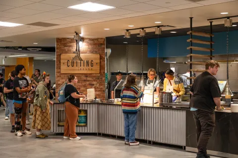 Students stand in line at a busy food counter marked “Cutting Board” where cooks prepare meals under warm lights.