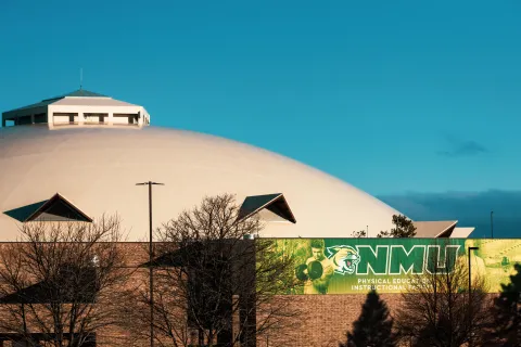 The Physical Education Instructional Facility is a two story brick building, with green NMU banners along its brick base. The dome is positioned behind the building under a clear blue sky. 