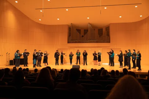 A vocal ensemble performs on a warmly lit concert-hall stage, and audience silhouettes in the foreground.