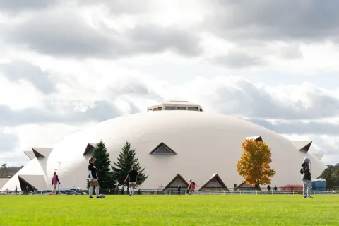 A large white domed arena stands behind a soccer field, where players stroll among small trees beneath a cloudy sky