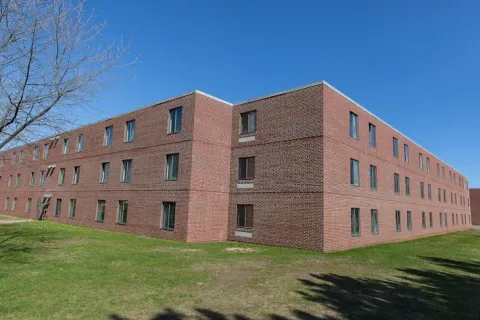 Exterior view of Spalding Hall, a large three-story red-brick building with rows of narrow windows stands on a green lawn beneath a clear blue sky.