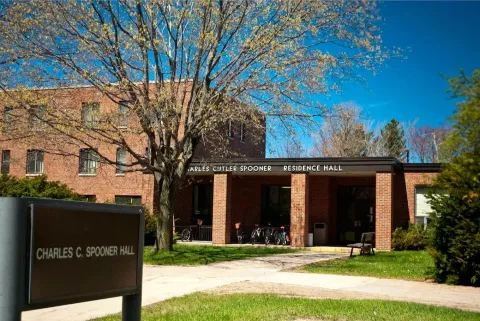A leafy tree shades the brick Charles C. Spooner Residence Hall, with its entrance, campus sign, bicycles, and sunny lawn beneath a clear blue sky.