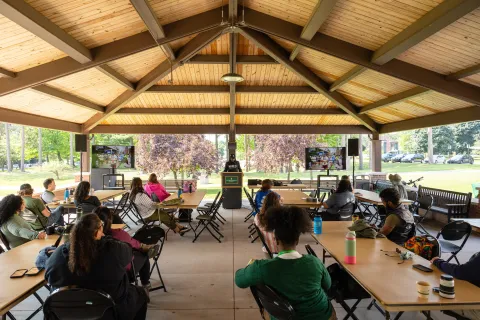 People sit at long tables in an open-sided wooden pavilion, listening to a speaker.