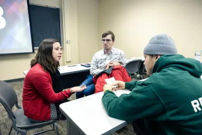 A woman in a red sweater talks to two men seated at a classroom desk; one man wears glasses and a plaid shirt, while the other wears a green hoodie and gray beanie. They appear to be having a serious discussion.