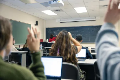 Students sit at desks with laptops open, raising their hands in a classroom while a teacher writes on a chalkboard at the front of the room. The classroom has fluorescent lights and a ceiling-mounted projector.