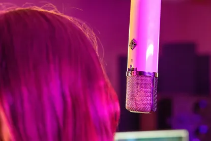 A close-up shot of a vintage-style microphone hanging in a recording studio, with a person with long hair, illuminated by pink and purple lighting, standing nearby.