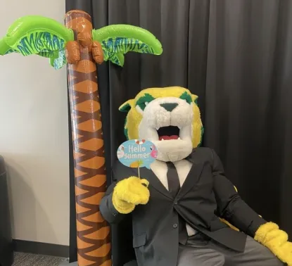 Wildcat Willy in a business suit, sitting next to an inflatable palm tree and holding a sign that says "Hello Summer!" 