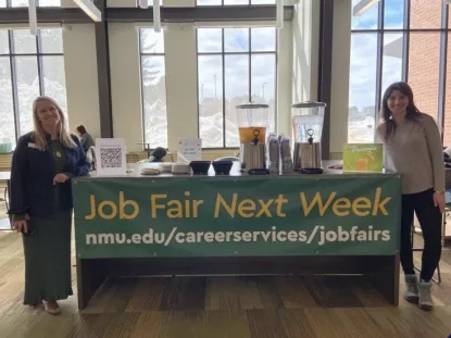 Katie Korpi, Director of Career Services, and Bia Guth Roque, one of the principal secretaries, stand beside a table filled with refreshments and a banner that says "Job Fair Next Week" 