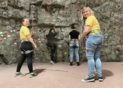 Two Career Services student employees wearing yellow shirts & climbing harnesses smile at the camera while helping two students climb an indoor rock wall