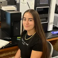 A young woman with long brown hair sits at a desk in a science lab, smiling at the camera. She is wearing a black shirt and tan pants. There is lab equipment, a computer, and an open notebook on the desk.