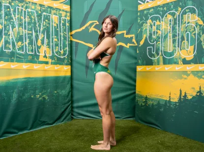 A photo of a collegate swimmer, her arms are crossed and she looks confidently over sher shoulder at us. She is dressed in NMU green