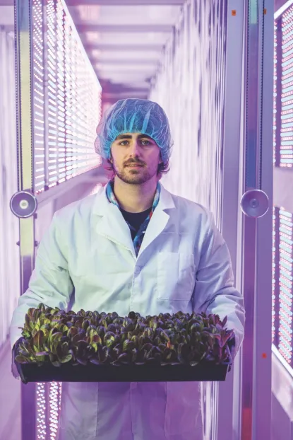 Young man in lab coat and hair net holding tray of leafy greens in indoor lab