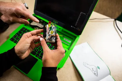 Hands holding small electrical device over bright green laptop computer with teacher's hand pointing