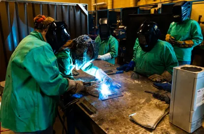 Five females in welding masks and green protective jackets watching two people welding on a worktable in a classroom lab
