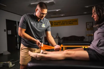 Woman sitting on medical table as man places an orange stretch band around her foot for physical therapy