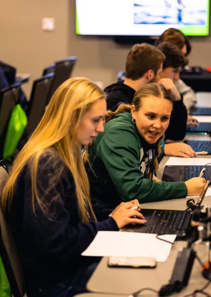 Multiple students at tables with computers working together to solve cyber security challenges
