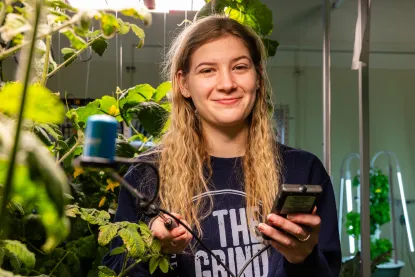 Young woman surrounded by tall plants indoors holding monitoring equipment