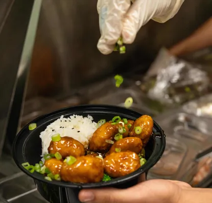 Gloved hand sprinkling green onion on an order of General's Chicken