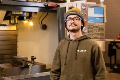 2026 Graduate, Josh Guinazzo, poses in front of a CNC machine in the manufacturing lab of the NMU Jacobetti Complex