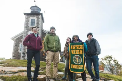 A group of students and faculty, holding an NMU banner, in front of a lighthouse
