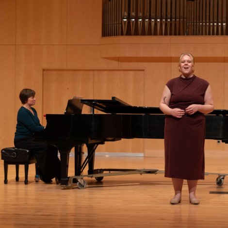 A singer in a burgundy dress performs beside a grand piano, with a pianist behind her in a warm, wood-paneled concert hall.