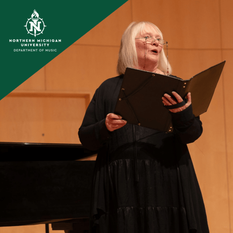 Northern Michigan University music recital: a woman sings from a black folder beside a piano on a warm-lit stage, conveying a focused, expressive mood.