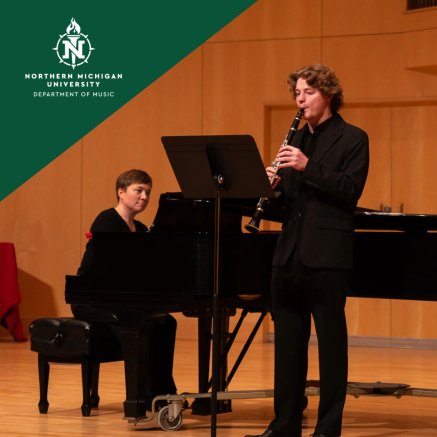 Clarinetist performs beside a pianist at a grand piano in a warm wood-paneled concert hall with organ pipes.