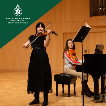 Northern Michigan University music performance: violinist in black plays beside a smiling pianist in an orange scarf on a concert hall stage.