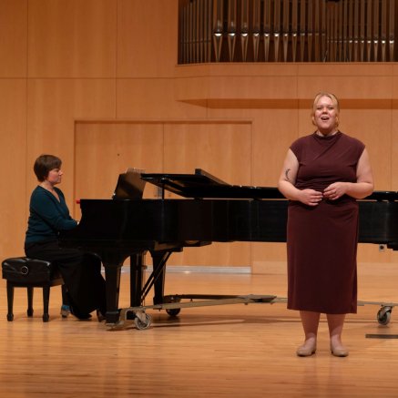 A singer in a burgundy dress performs beside a grand piano, with a pianist behind her in a warm, wood-paneled concert hall.