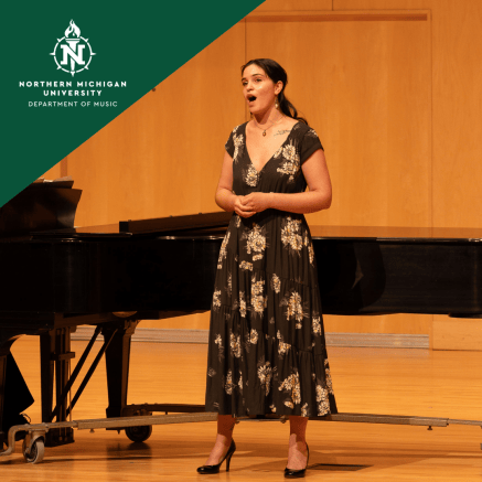Female vocalist in a black floral dress sings beside a grand piano in a wood-paneled recital hall; Northern Michigan University logo appears.