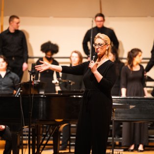 Choir director Dr. Erin Colwitz addressing the audience, choir students and accompanist in background