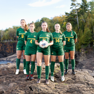 five female soccer players standing on the shore of lake superior