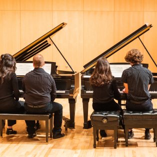 Two pairs of painists sit at two grand pianos, backs facing the viewer