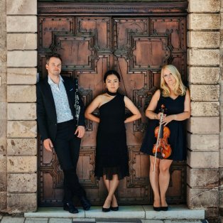 Man in black suit with light blue shirt holds clarinet, pianist stands with arms and legs crossed, and woman with blonde hair holds a violin at her front in front of a large, fancy wooden door.