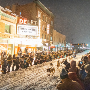 A crowd lines the snow covered street. Downtown Marquette businesses are lit up. A sled dog team races down the middle of the road. 