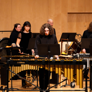 student with medium length curly blonde hair dressed in all black plays a marimba in front, with other students also in all black playing other instruments in the background